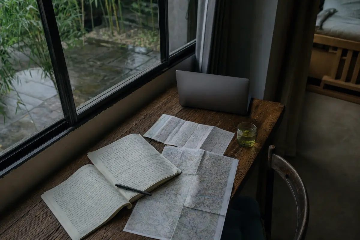 A desk with an open notebook, a laptop, a folded map and a cup of tea beside a window