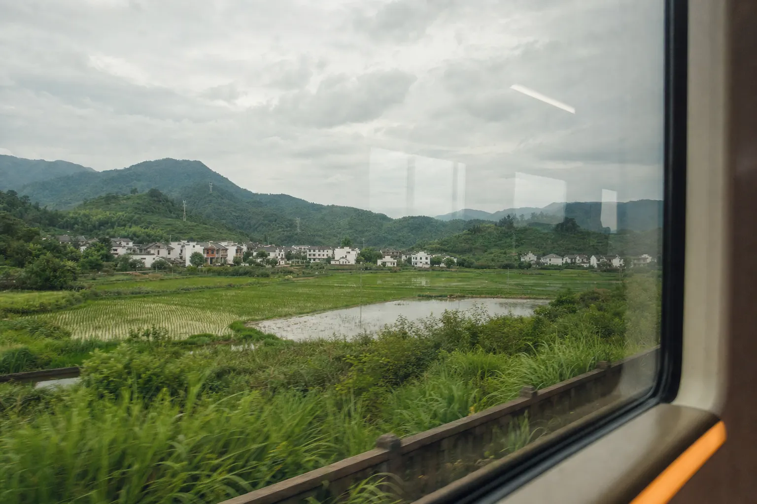 Rice paddies, a white walled village and low green hills seen from a moving train window