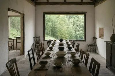A long wooden table laid with plain ceramic bowls in a whitewashed farmhouse room, bamboo forest through the window