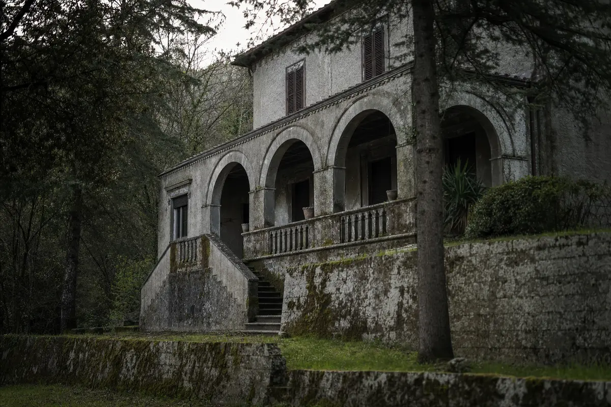 A grey stone villa with an arched veranda and weathered shutters standing among trees