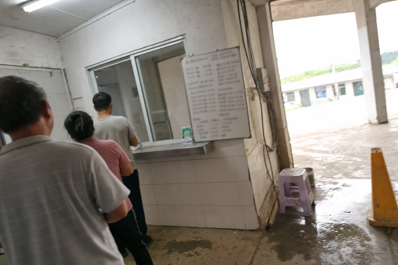 A ticket window with a hatch and a handwritten price board in Chinese, seen at an angle with a queue of people from behind