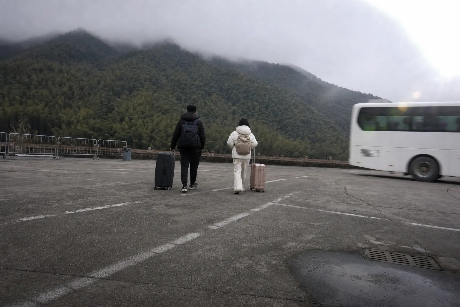 Two people with wheeled cases seen from behind on a paved forecourt, a coach pulling away, bamboo hills in cloud beyond