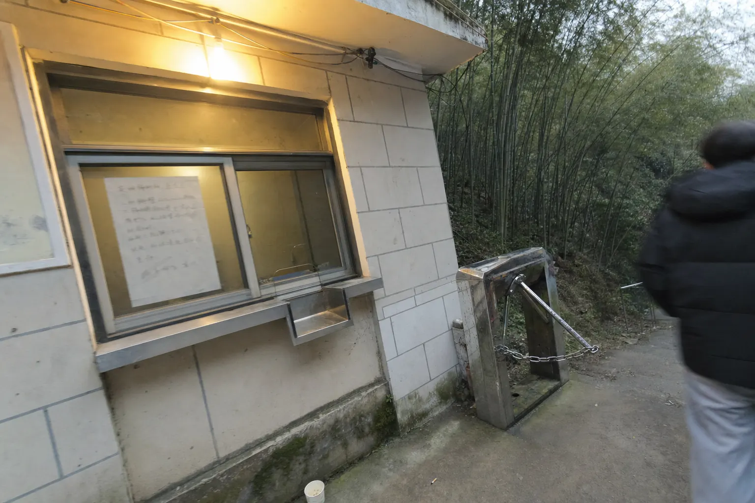 A hillside ticket kiosk with a metal turnstile beside it, bamboo rising behind and a handwritten notice taped to the glass