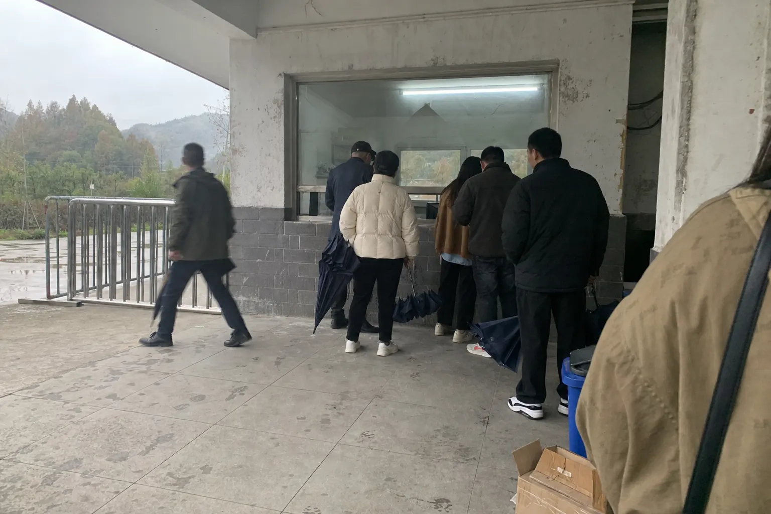 A short queue at a ticket window under a concrete canopy, furled umbrellas in hand, the light flat and grey