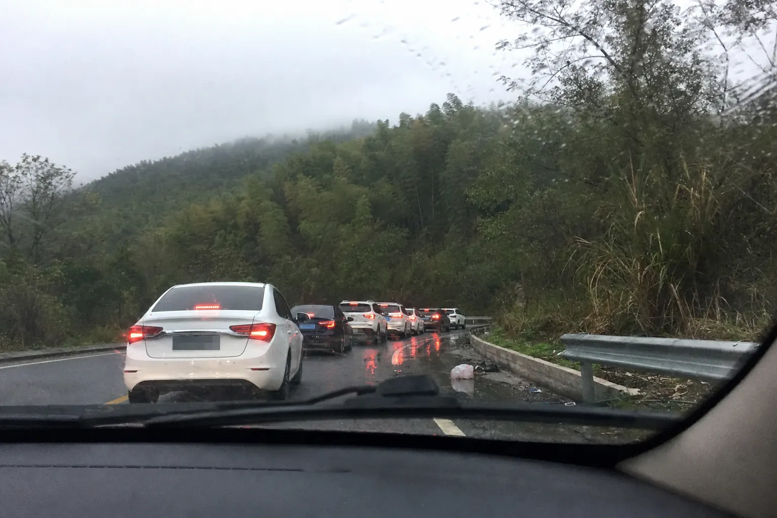 A queue of cars backed up on a wet county road below a bamboo ridge, seen through a rain flecked windscreen