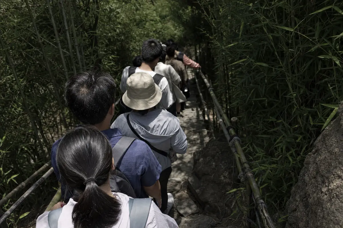 A queue of visitors packed onto a narrow stone mountain path in harsh summer light