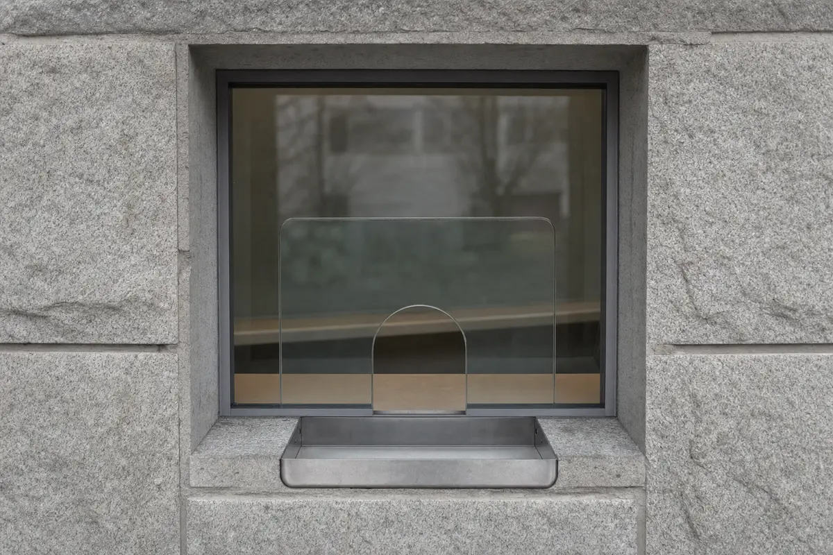 A small ticket window with a glass hatch and metal tray in a stone kiosk