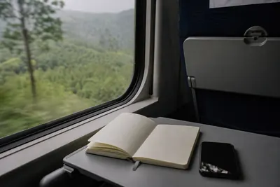 An open planning notebook and a phone timetable on a train tray table, hills passing outside