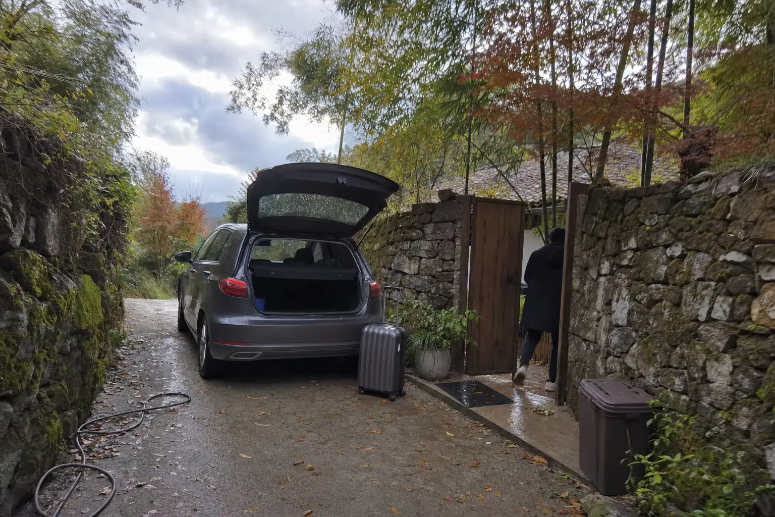 A parked car with a suitcase beside it on a narrow lane between stone walls and bamboo, a guesthouse gate half open