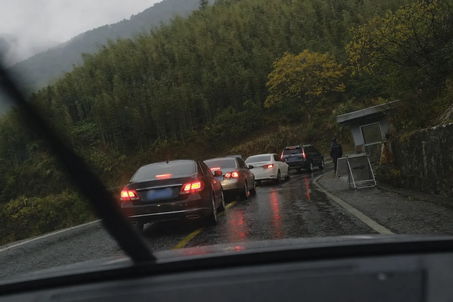 A queue of cars on a wet mountain road under bamboo, brake lights on, with a turn around sign ahead