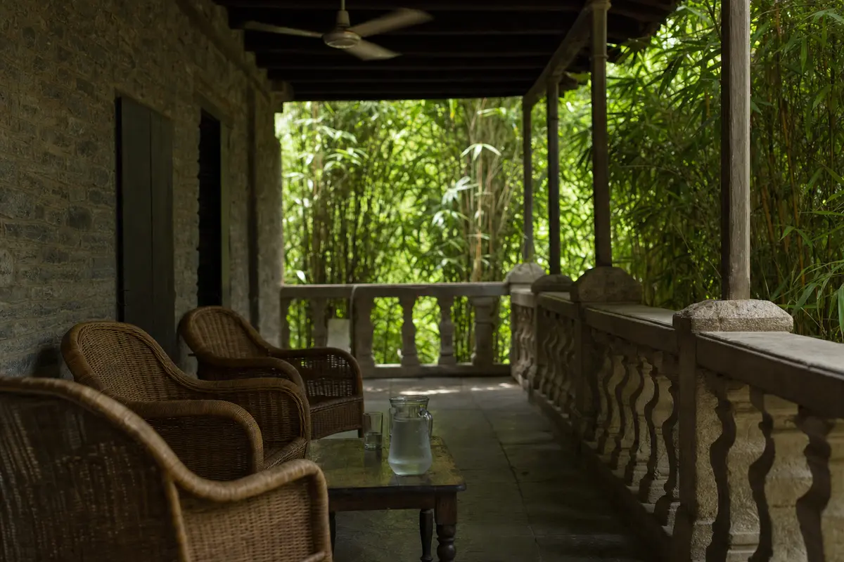 A shaded guesthouse terrace with wicker chairs and a ceiling fan above green bamboo