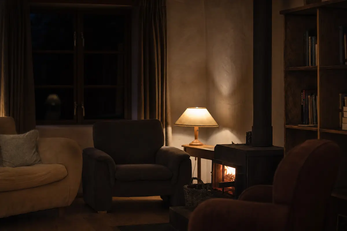 A guesthouse common room in the evening with low lamps, a wood stove and empty chairs