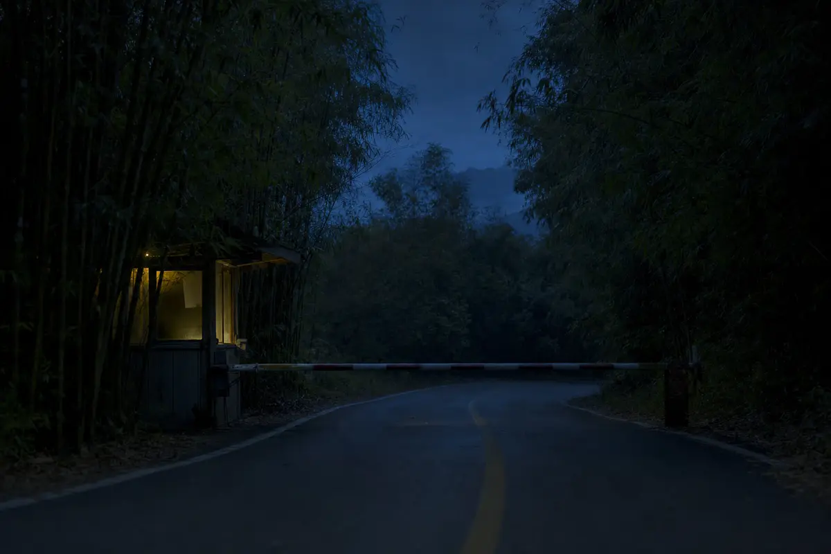 A road barrier down across a mountain road at dusk with one light on in the hut