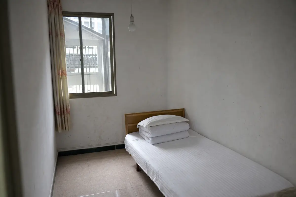 A plain guesthouse bedroom with a narrow bed, a tiled floor and a bare pendant bulb