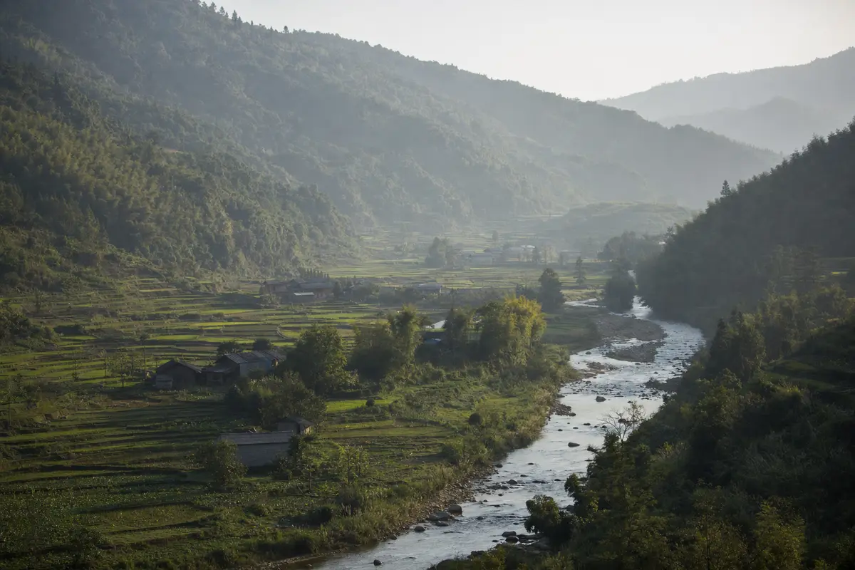 A broad green valley with terraced fields and scattered buildings below forested ridges