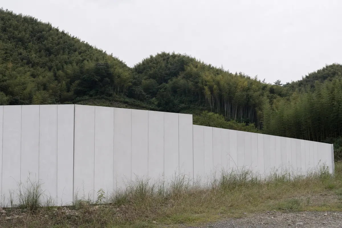 A plain unmarked construction hoarding around an empty rural site below bamboo hills