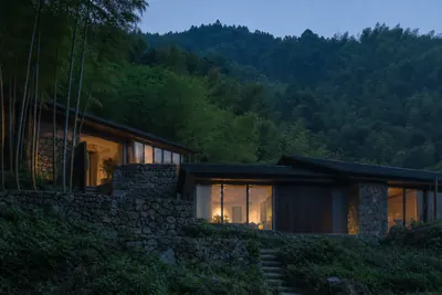 A low stone and timber guesthouse lit from inside at dusk, set into a bamboo hillside
