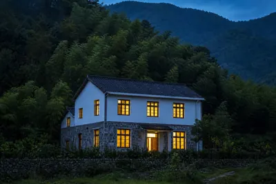 A whole two-storey stone and plaster house lit from inside at blue hour below a dark bamboo slope