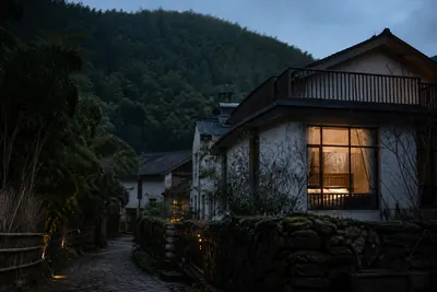 A lit guesthouse window on a village lane at dusk with bamboo hills standing behind it