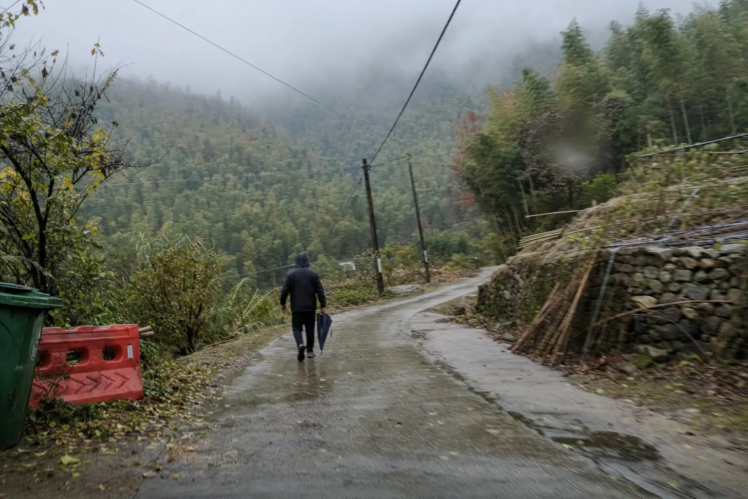 A wet concrete lane winding down through bamboo under low cloud, a walker in a dark hooded jacket seen from behind carrying a closed umbrella, a red plastic barrier and a green bin at the roadside
