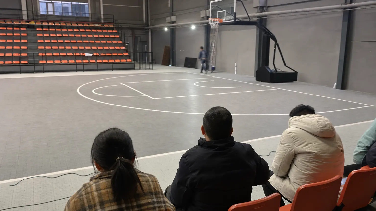 Spectators seen from behind taking their seats beside an indoor three on three basketball court under uneven arena lighting