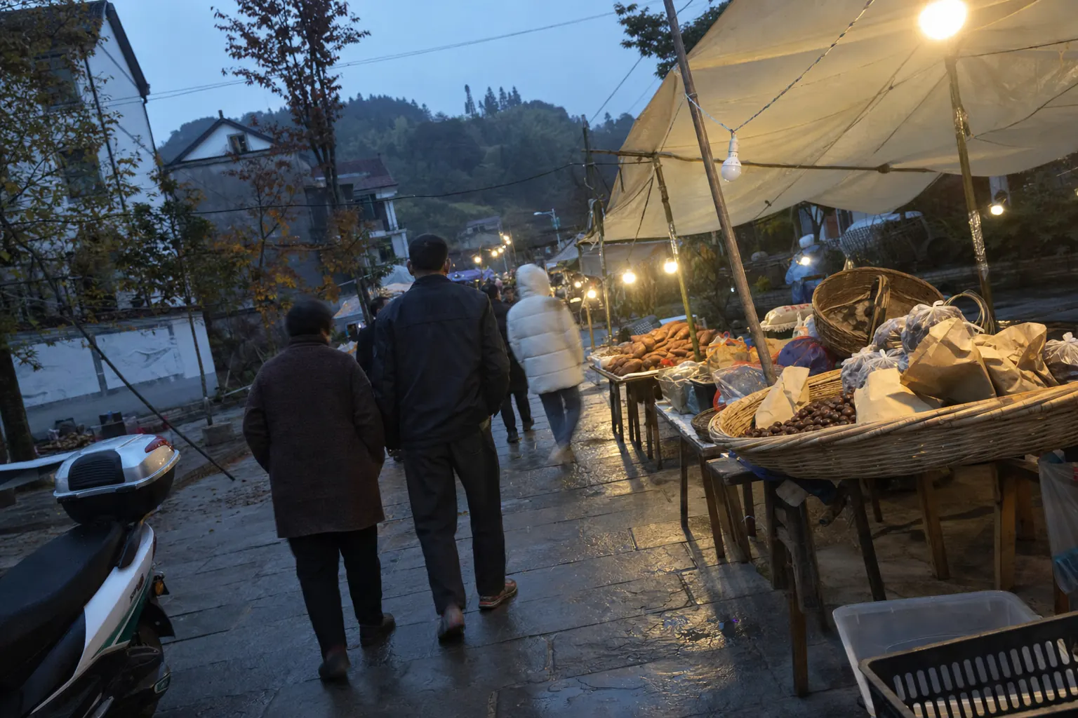 People walking away down a wet stone village street at dusk, past trestle tables of sweet potatoes and a wide bamboo basket of chestnuts under a sagging tarpaulin and bare bulbs, a scooter cut off at the left and a wooded hill behind