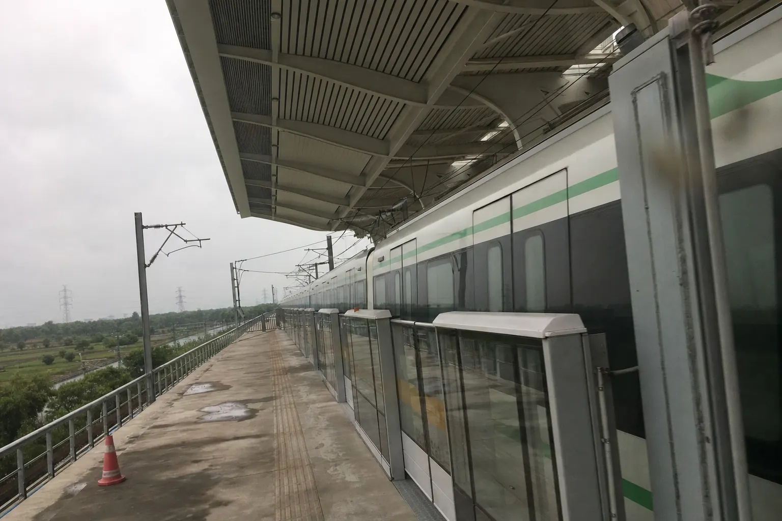 A pale new metro train standing at an empty elevated platform under a grey sky, the platform doors shut and a wet patch on the concrete