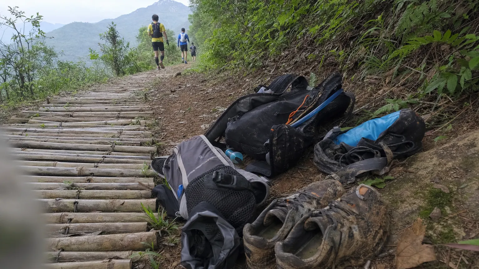 A row of trail running packs and muddy shoes beside a bamboo path in flat morning light, with runners visible only from behind in the distance