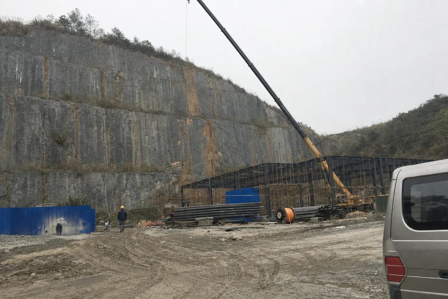 A stepped grey quarry face with a half built steel frame and scaffolding at its foot, a crane arm cutting diagonally across the rock, stacked pipes and churned mud in the foreground under a flat overcast sky