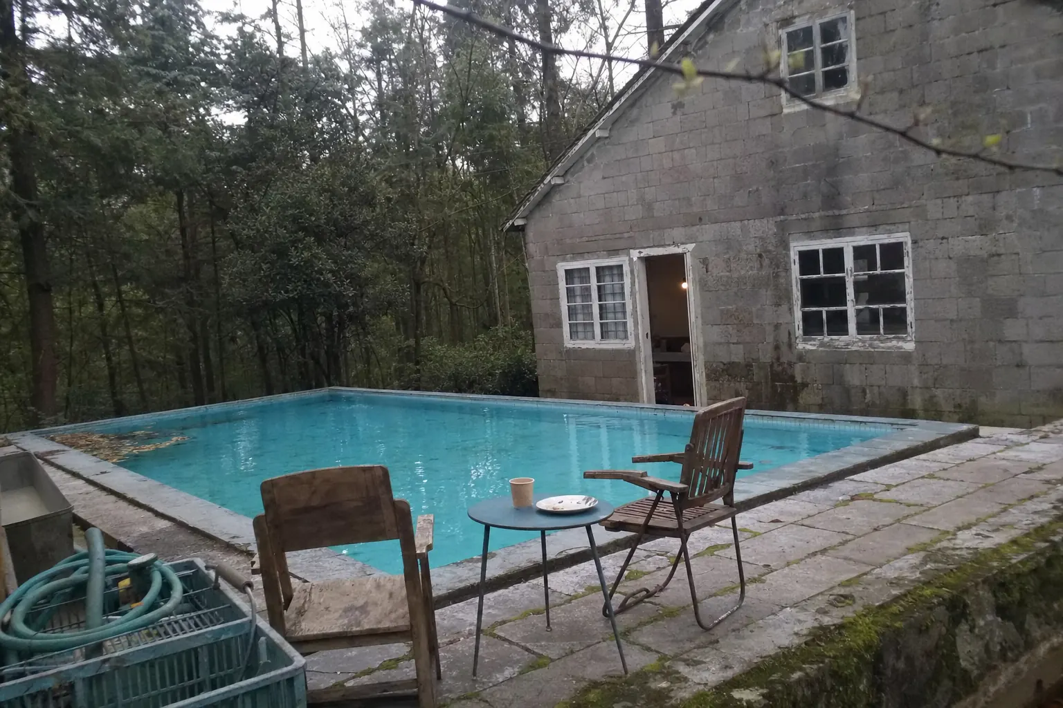 An old open air swimming pool full of clear water among tall trees, two mismatched wooden chairs and a paper coffee cup on a small metal table at the mossy stone edge, a grey stone building with peeling white window frames behind