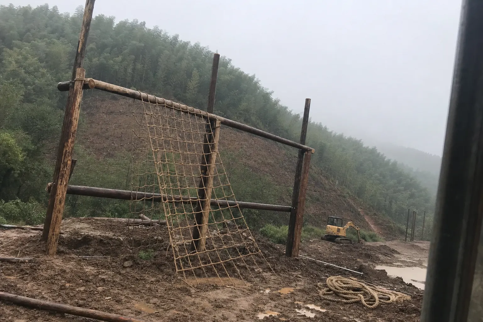 A rope net and timber obstacle frame half built on a muddy hillside clearing, a digger parked beyond it and bamboo on the ridge behind
