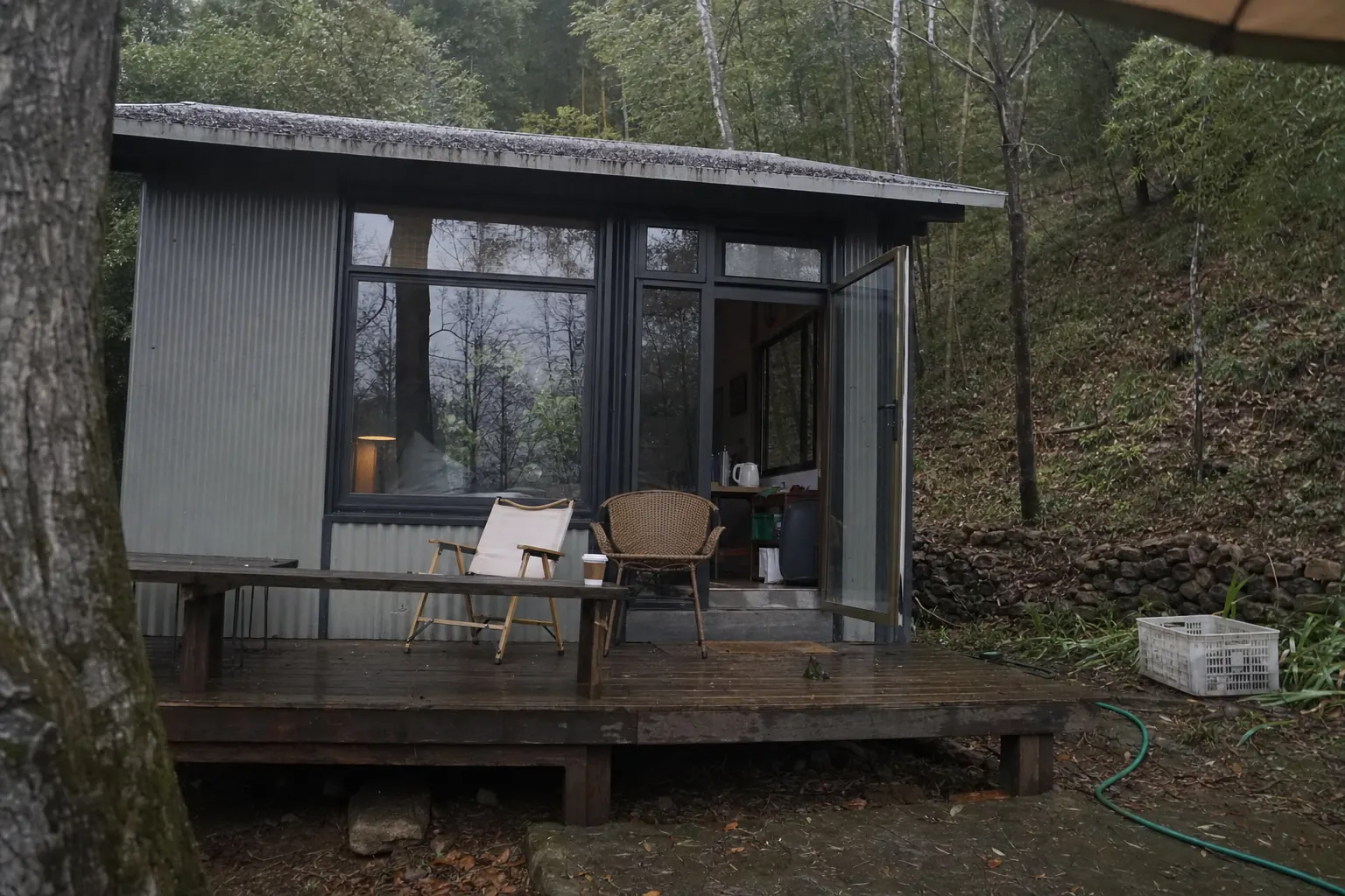 A small corrugated metal cabin with big windows among trees on a hillside, two mismatched chairs on its wet deck and a paper coffee cup left on the bench, overcast light