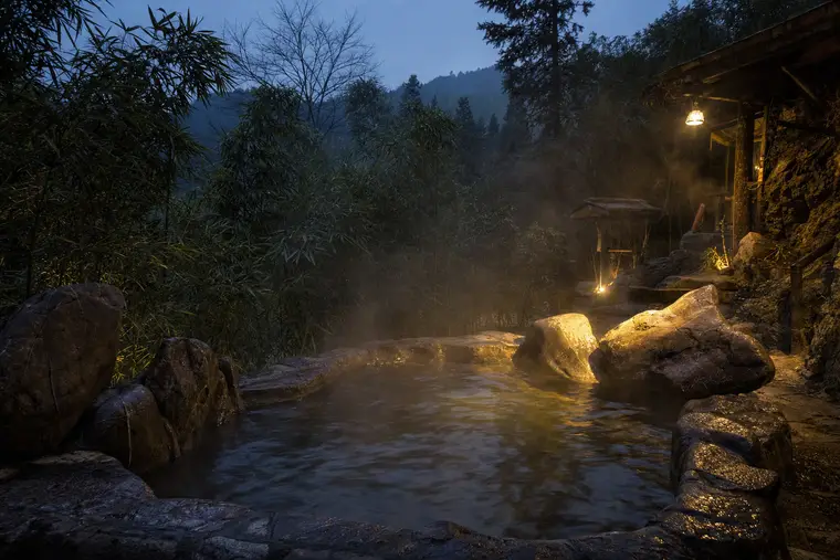 A steaming outdoor stone hot spring pool on a wooded hillside at dusk